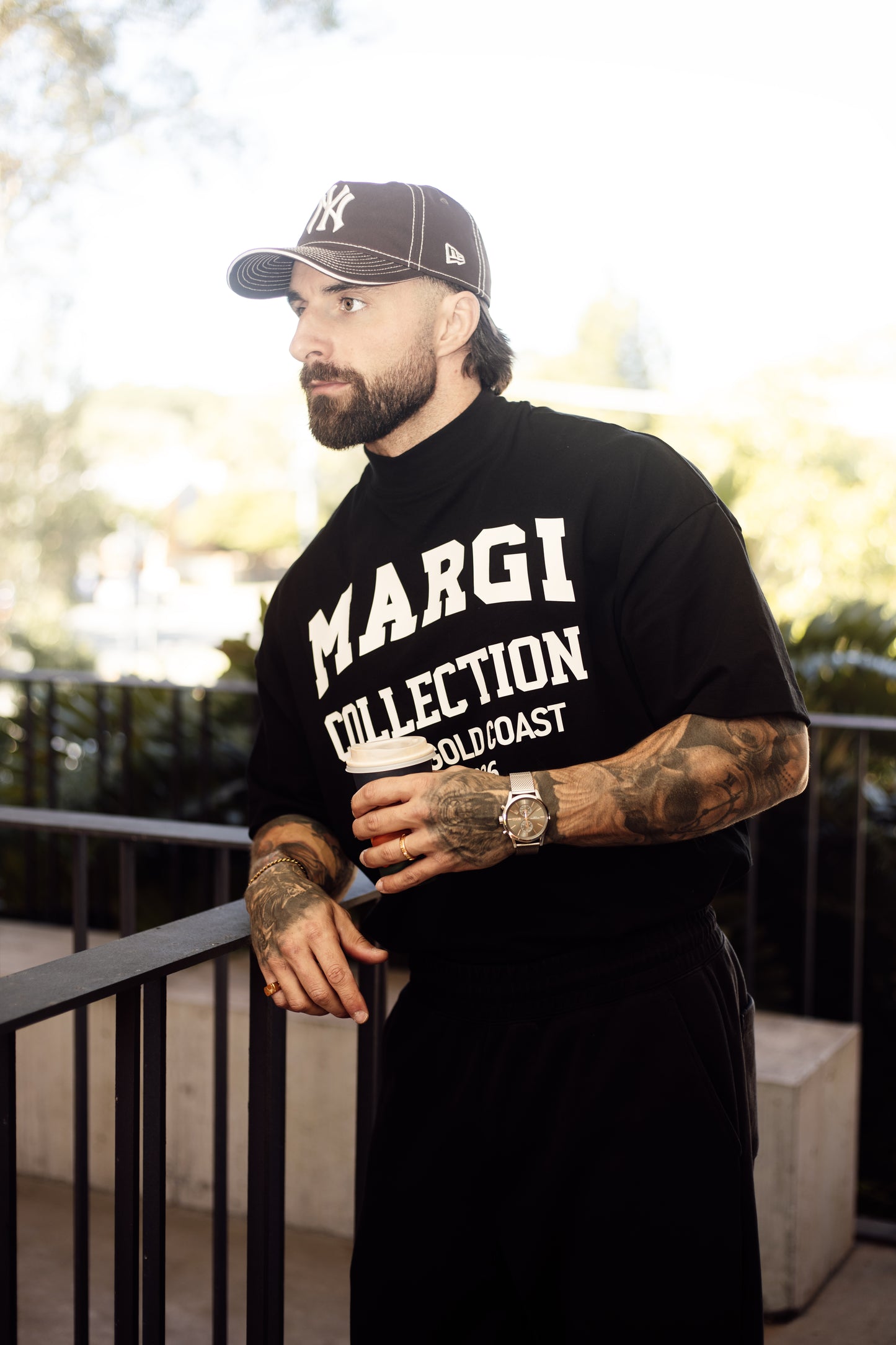 Man wearing a black t-shirt with 'MARGI COLLECTION' text, holding a drink on a balcony.