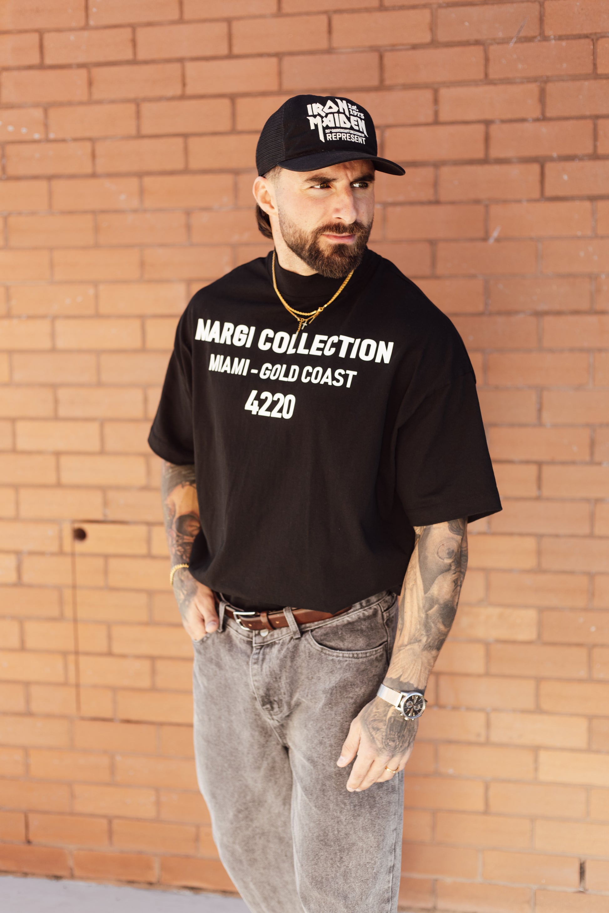 Man wearing a black t-shirt with text and a cap, standing against a brick wall.