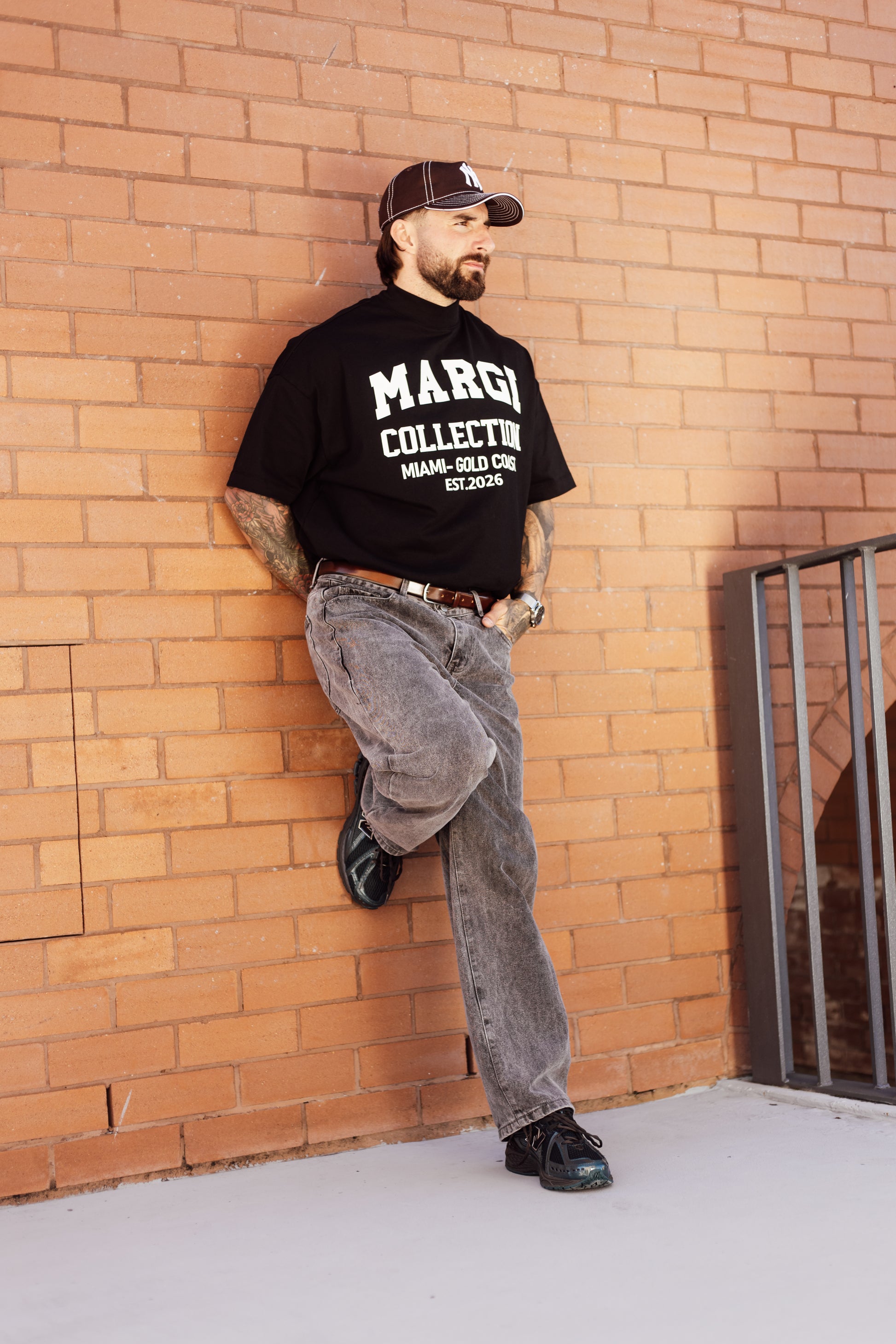 Man wearing a black t-shirt with 'MARGI' text, leaning against a brick wall.
