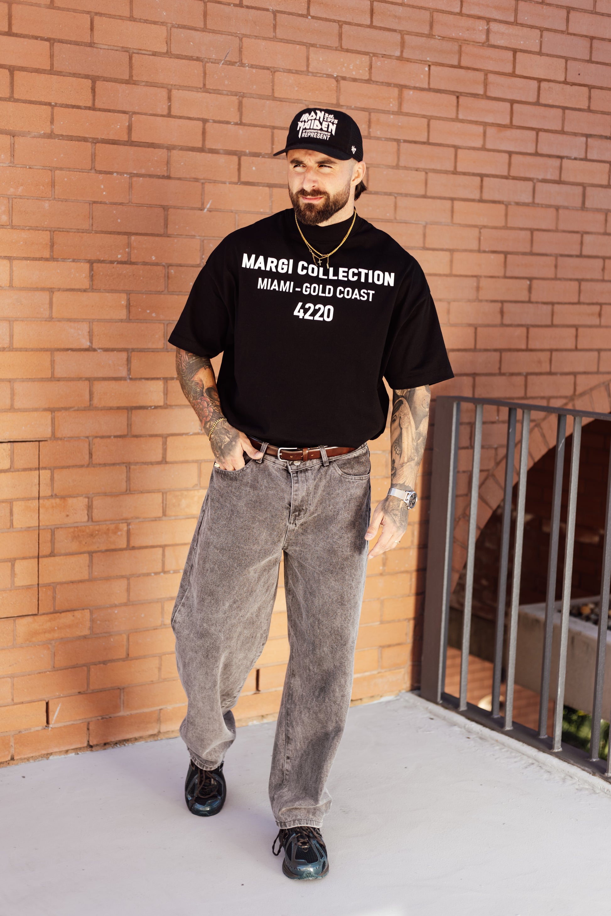 Man wearing a black t-shirt with text, gray pants, and a cap standing against a brick wall.
