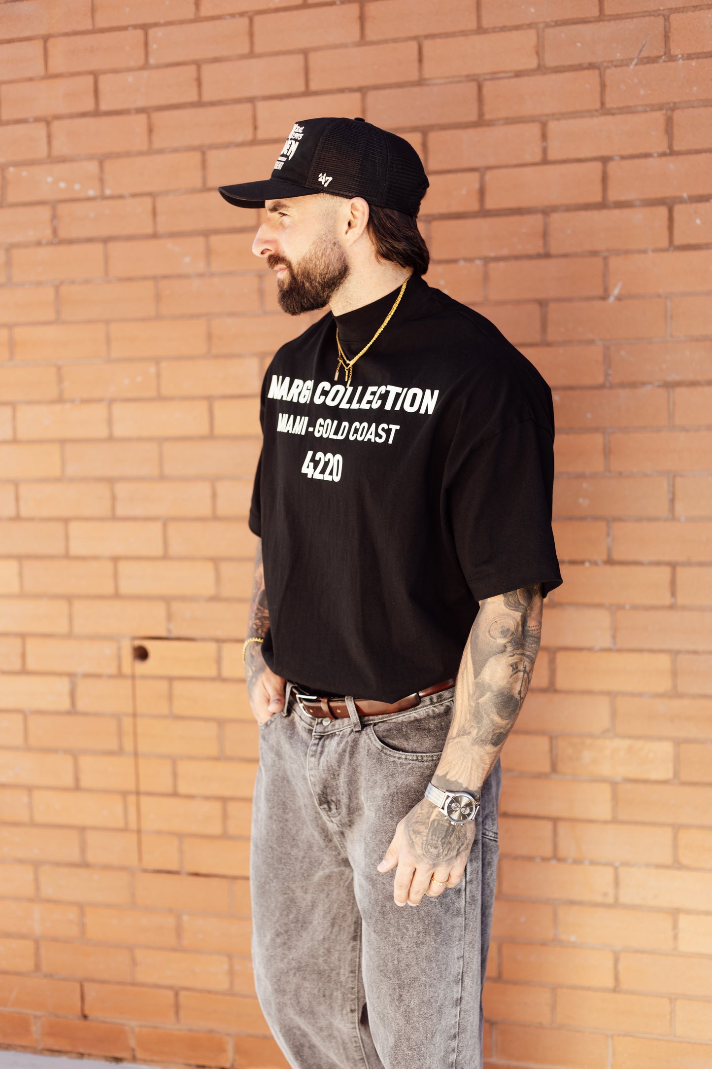 Man wearing a black t-shirt with text, cap, and gray pants standing against a brick wall.