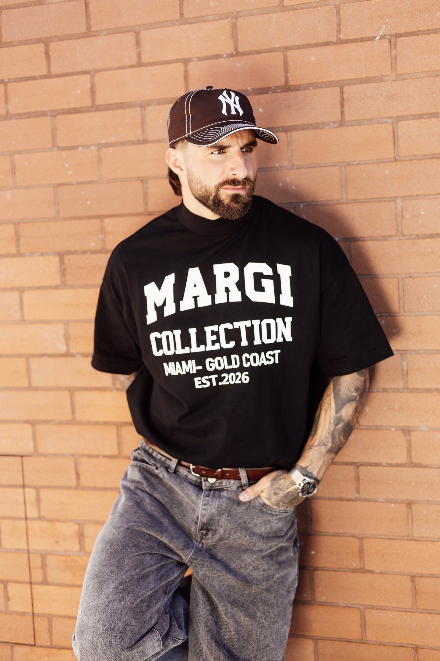 Man wearing a black 'MARGI COLLECTION' t-shirt and a cap against a brick wall.