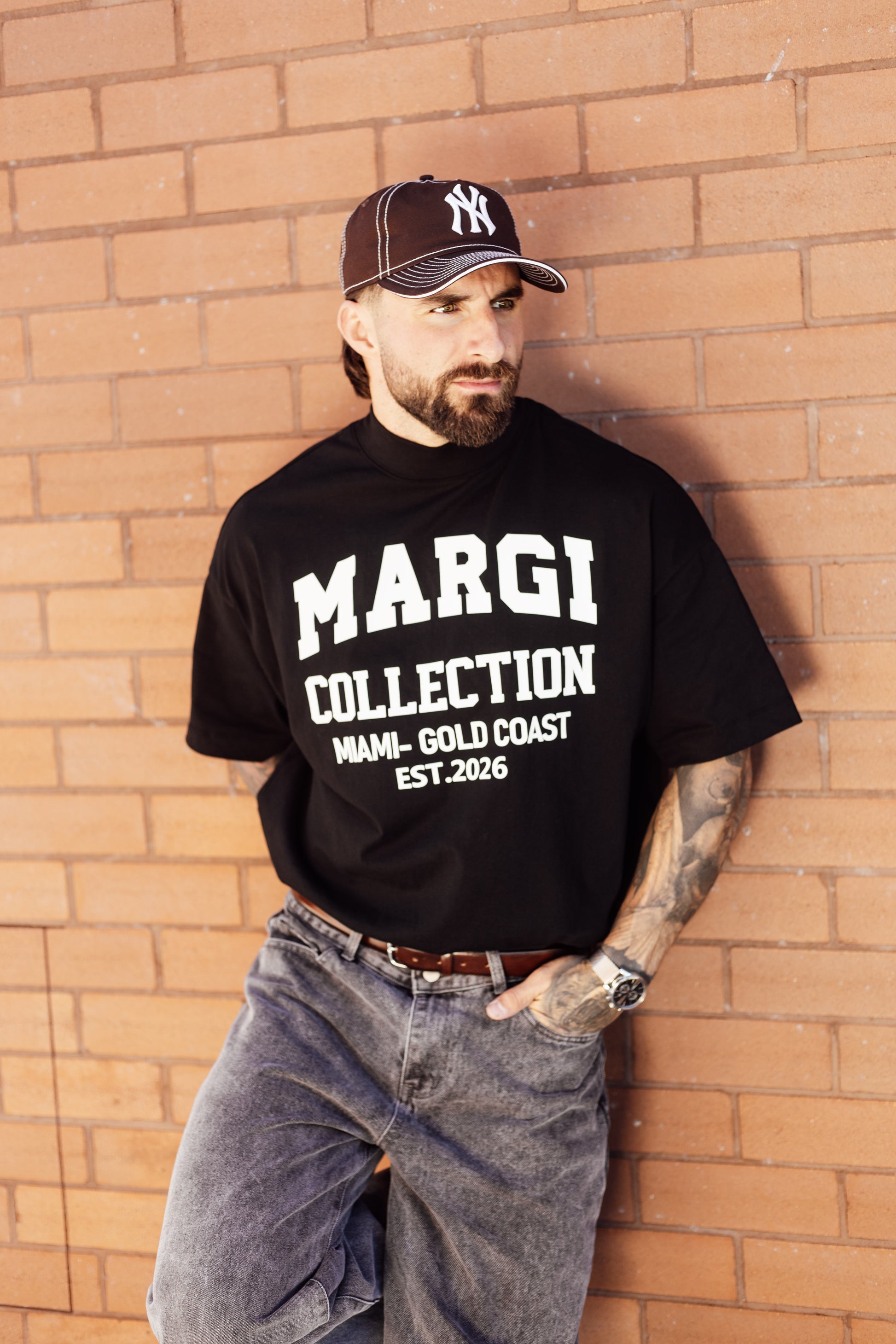 Man wearing a black 'MARGI COLLECTION' t-shirt and a cap against a brick wall.