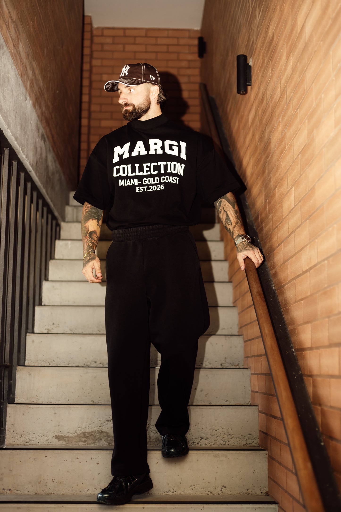 Man wearing a black 'MARGI COLLECTION' t-shirt standing on a staircase.