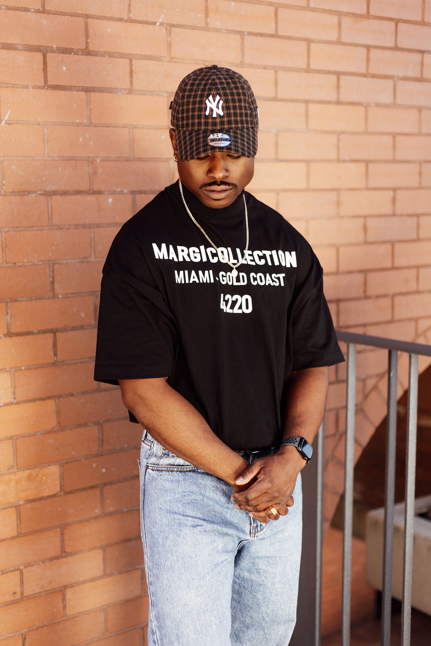 Man wearing a black t-shirt with text and a cap, standing against a brick wall.