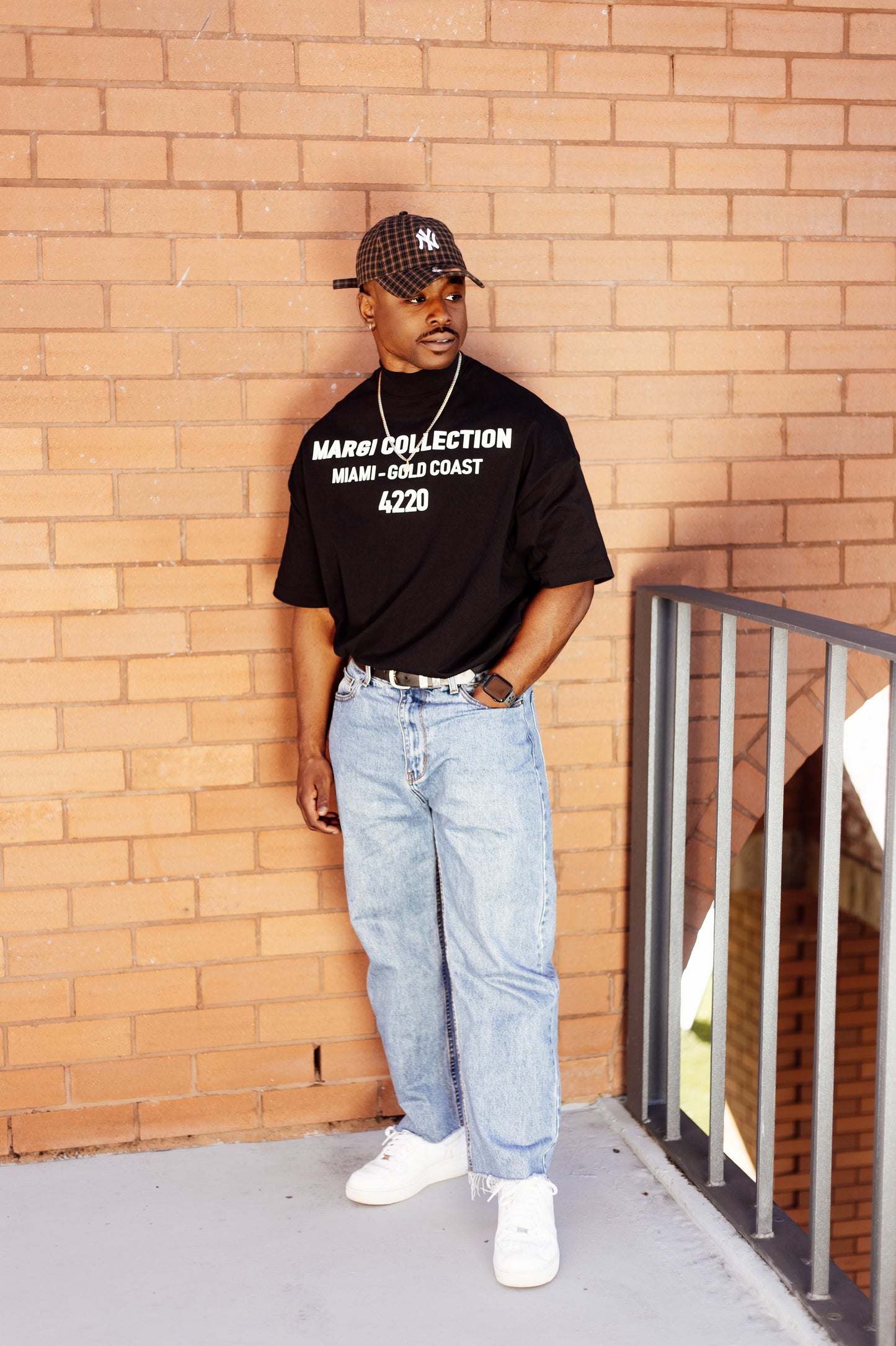 Man wearing a black t-shirt with text, blue jeans, and white sneakers leaning against a brick wall.