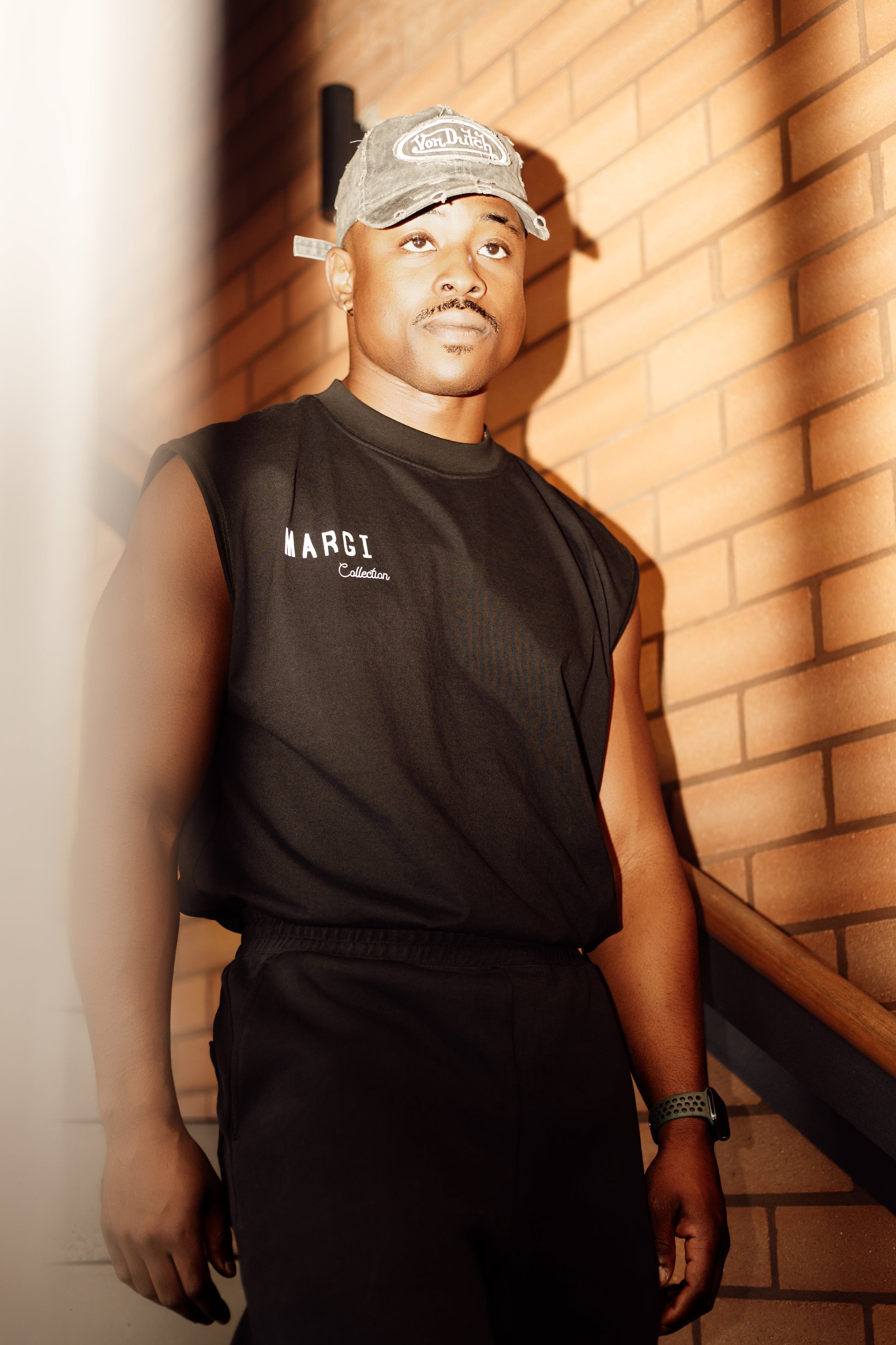 Man wearing a black sleeveless shirt and cap standing against a brick wall.