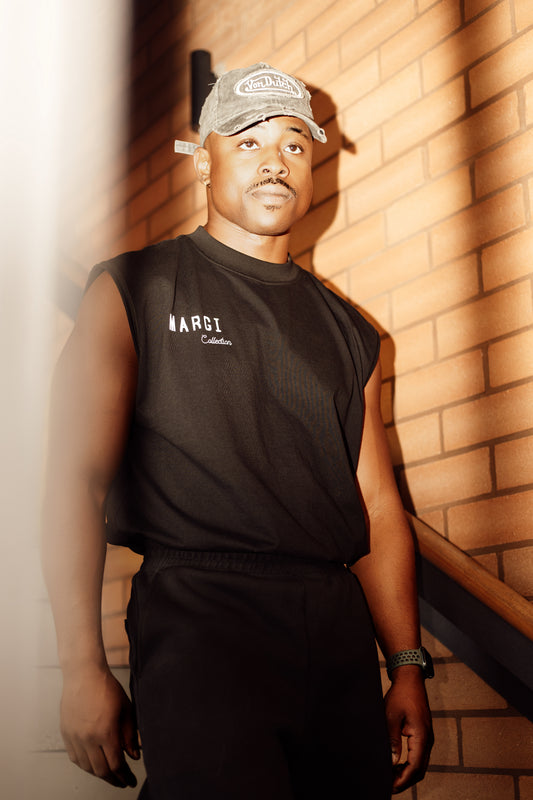 Man wearing a black sleeveless shirt and cap standing against a brick wall.