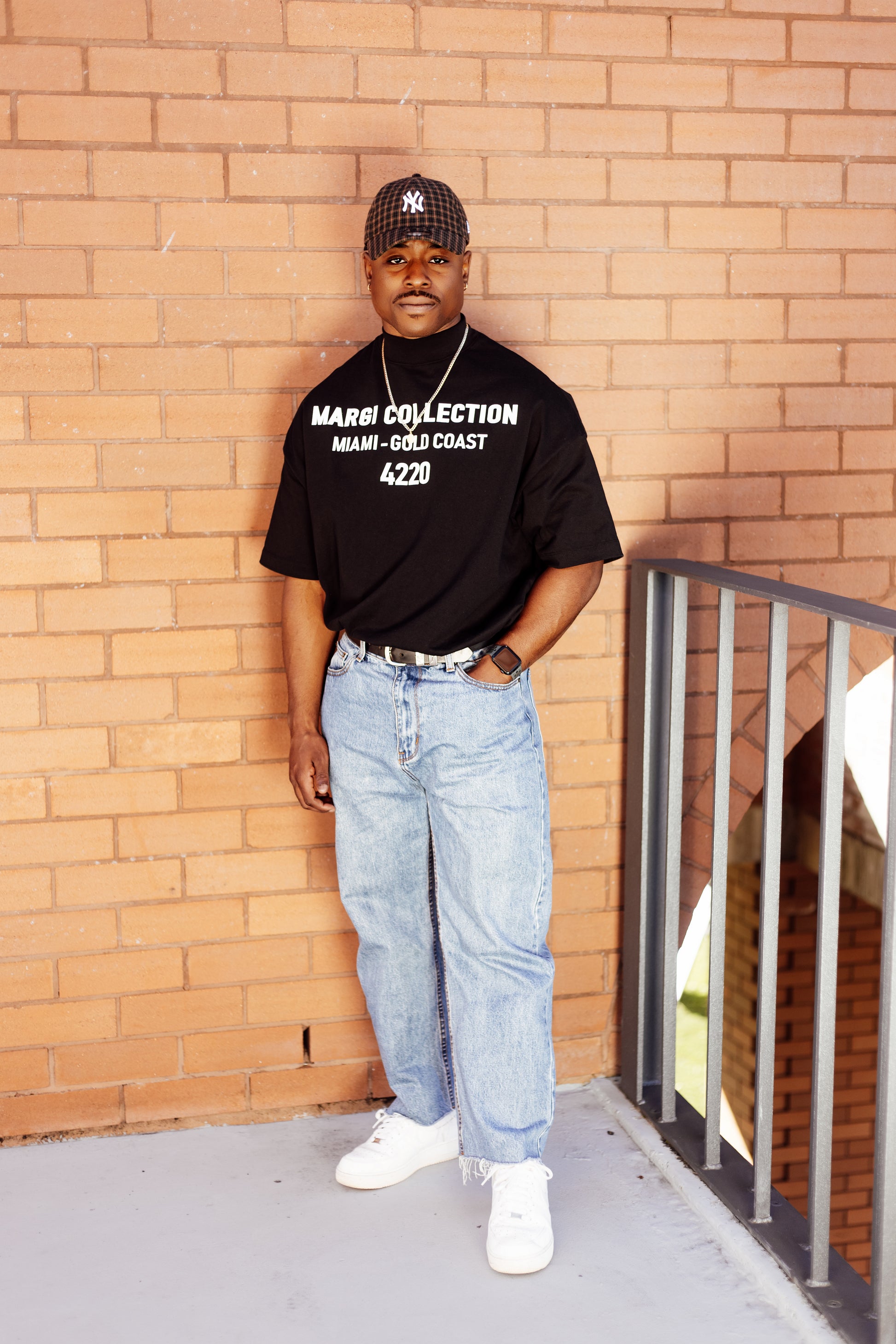 Man wearing a black t-shirt with text, blue jeans, and white sneakers against a brick wall.