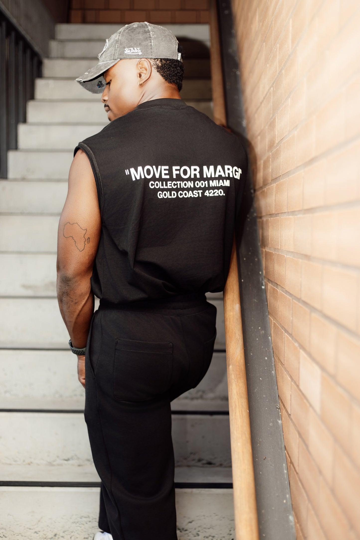 Person wearing a black sleeveless shirt with text, leaning against a wall on a staircase.