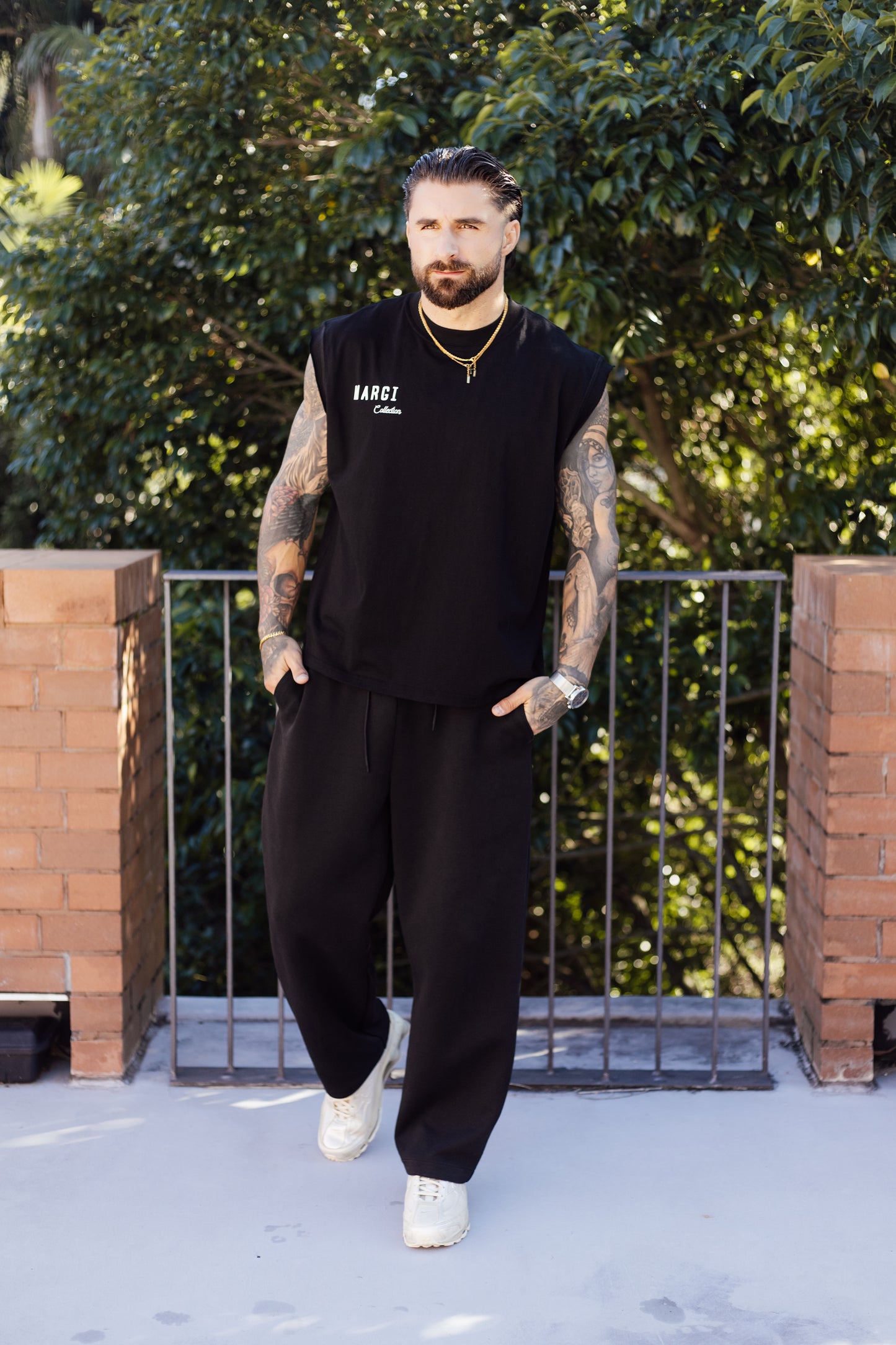 Man wearing a black sleeveless shirt and pants standing outdoors with greenery in the background