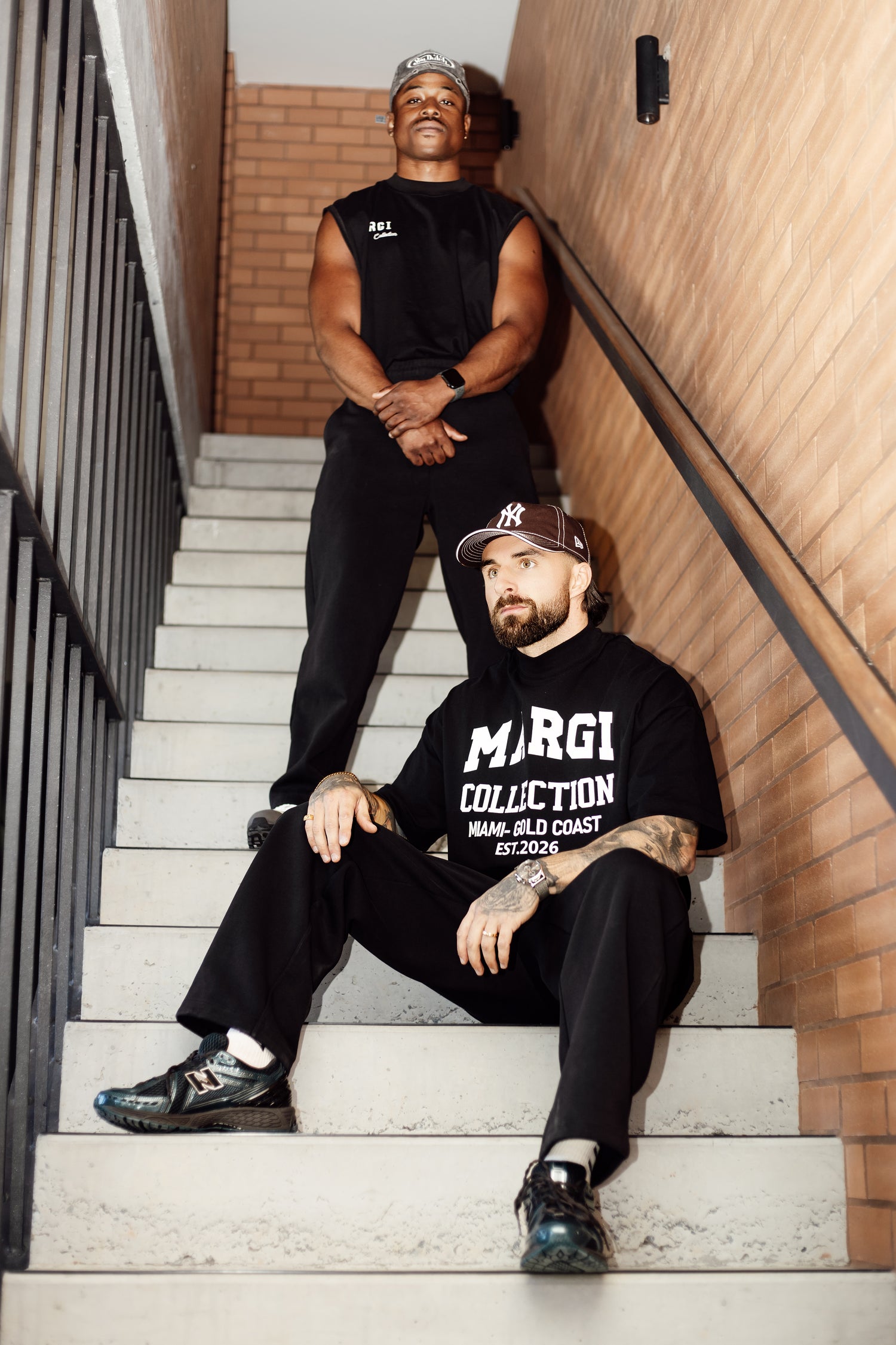 Two men sitting on a staircase, one wearing a black sleeveless shirt with visible branding and the other wearing a black t-shirt with 'MARGI' branding.